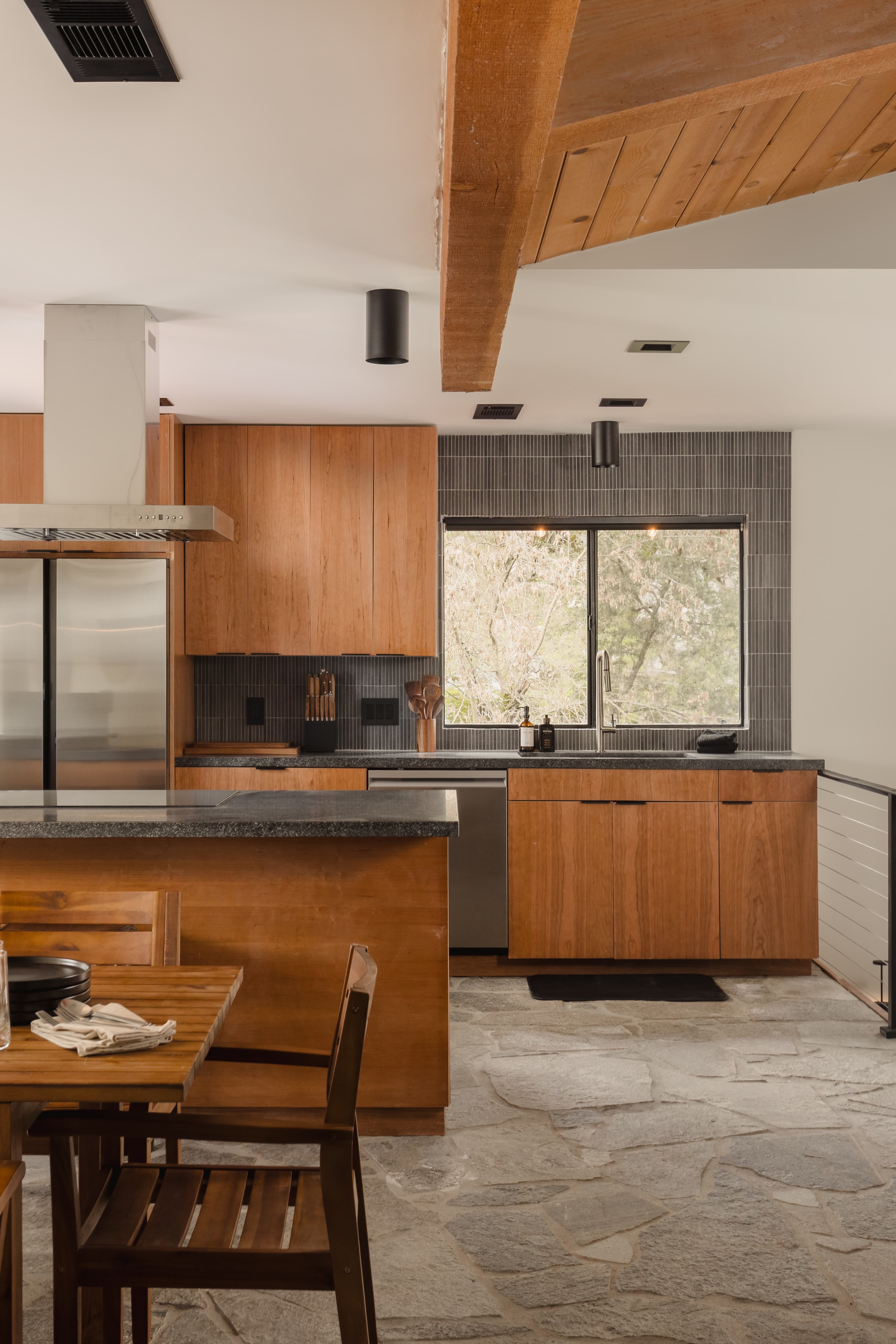 Kitchen with walnut cabinetry, vertical slate backsplash, granite island, stainless appliances, and a forest window beneath a vaulted cedar-beam ceiling — The Streamhouse